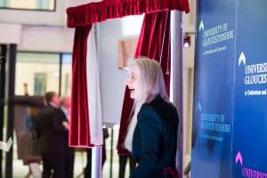 A woman smiles next to a brass plaque framed by red curtains.