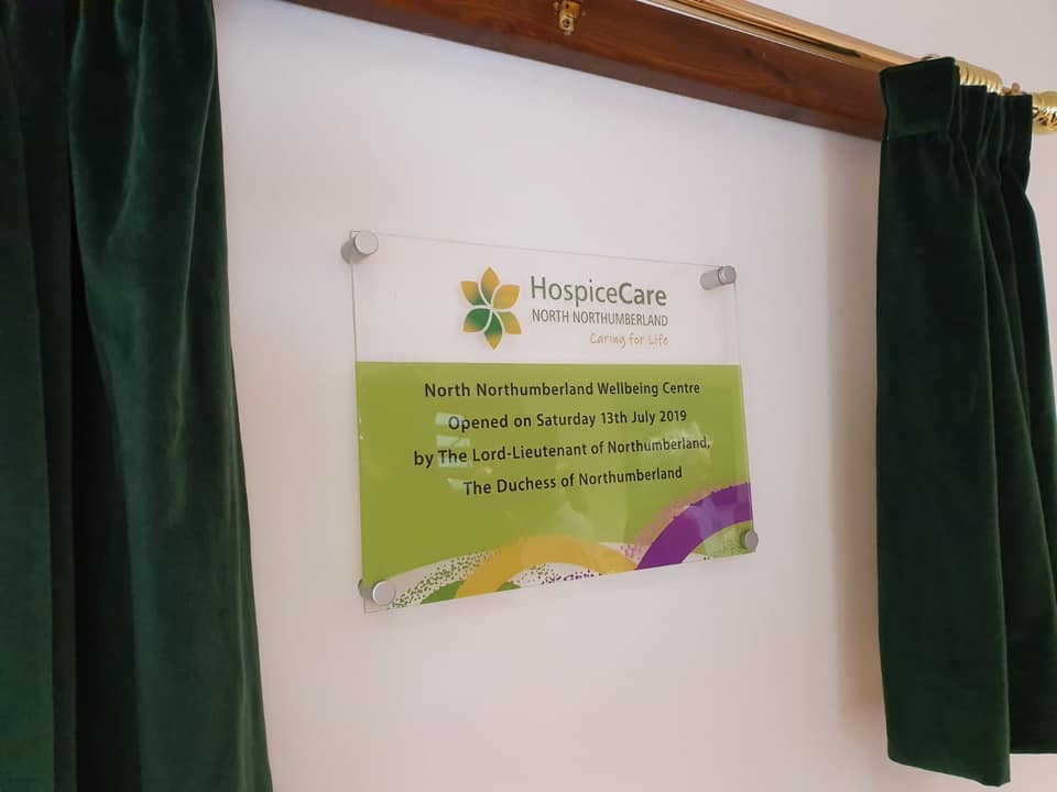 A colorful Perspex plaque framed by green curtains. It reads in part "HospiceCare North Northumberland, Caring for Life."
