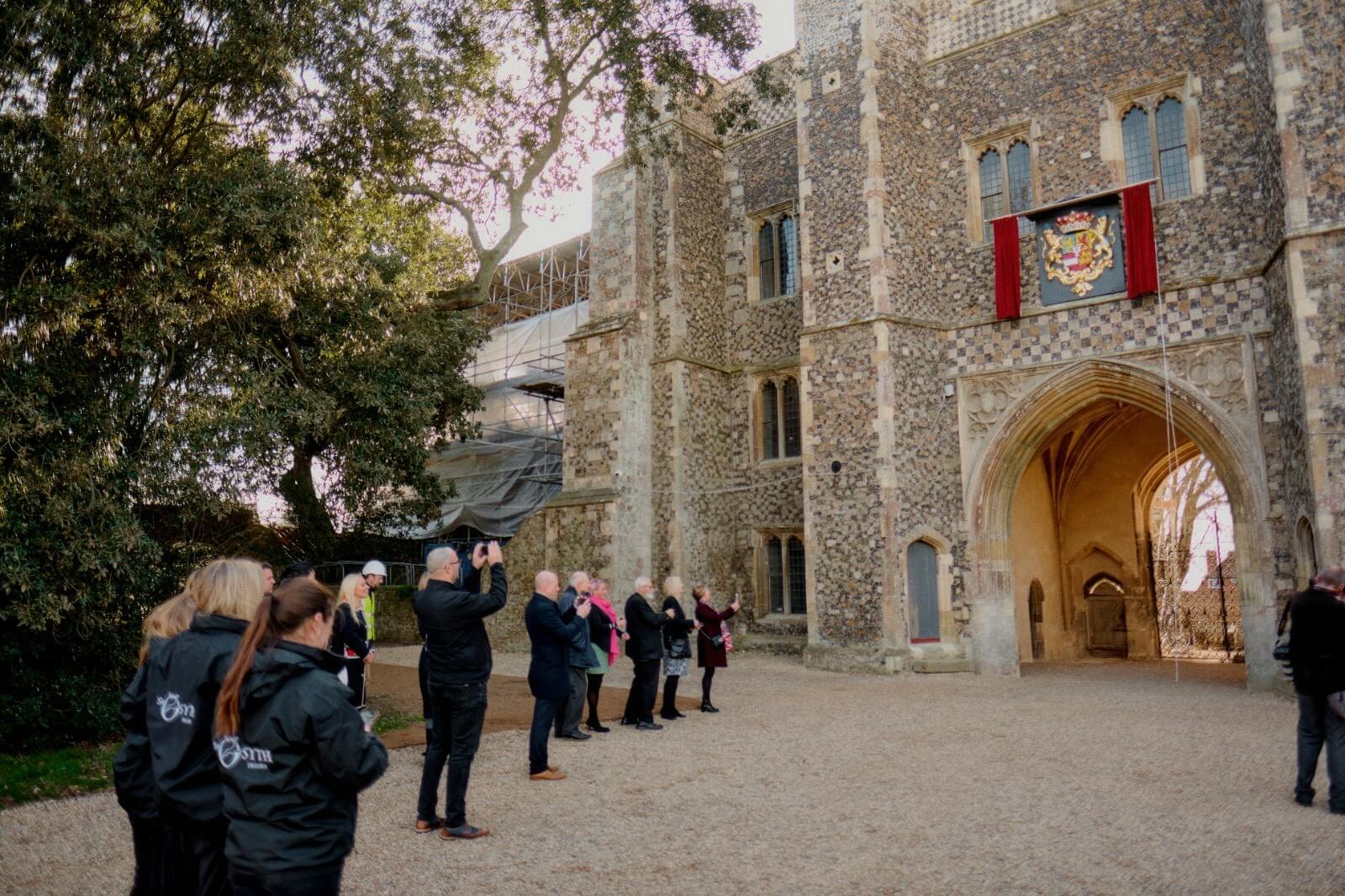 A group of people taking pictures of an old building that has a crest framed by red curtains.