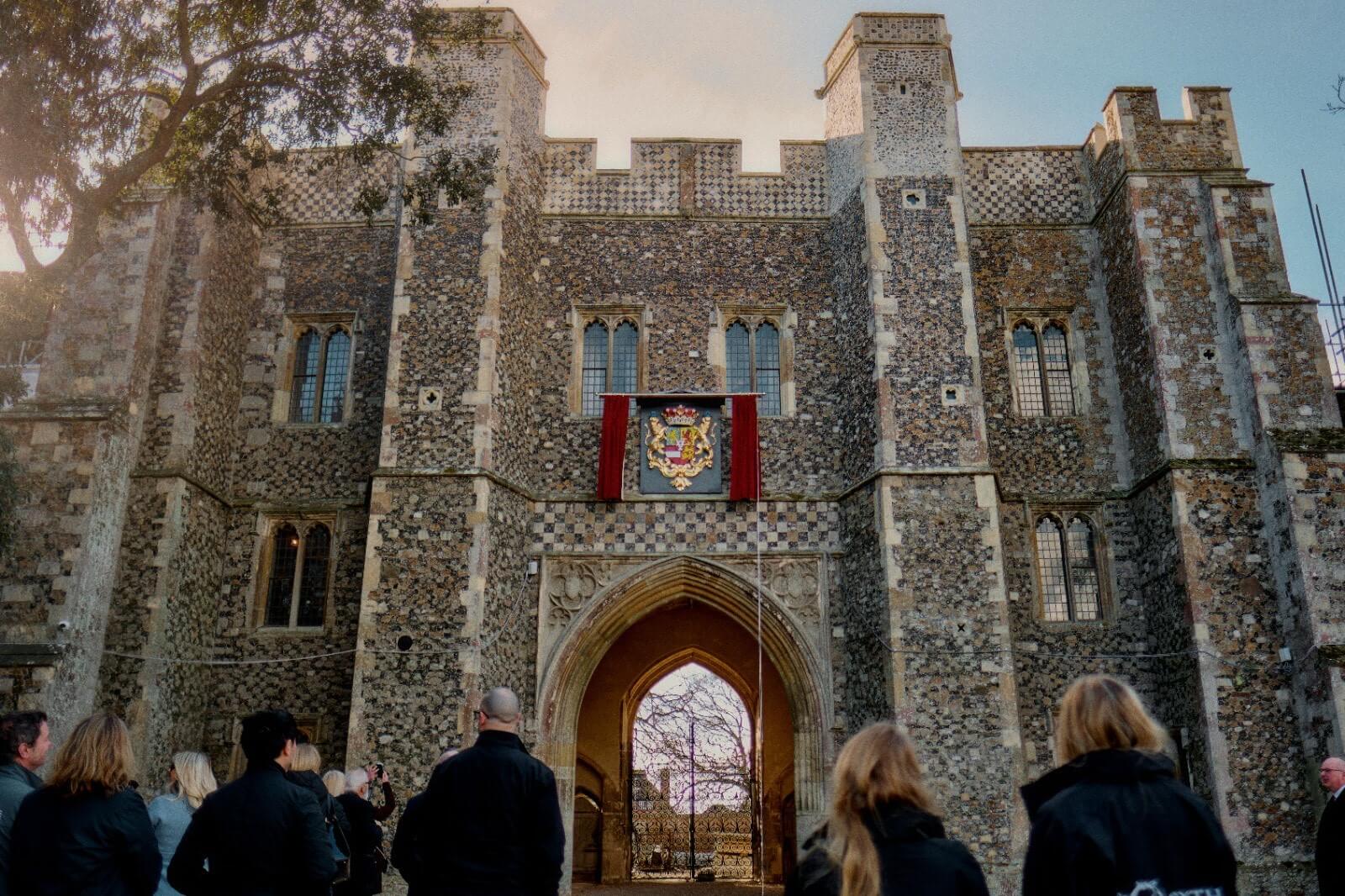 A wide shot of the old building with the crest framed by red curtains. A group of people faces the building, away from the camera.