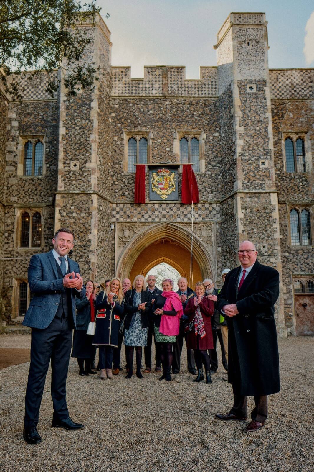 A crowd of people standing in front of an old building. Over the arch is a crest framed by red curtains.