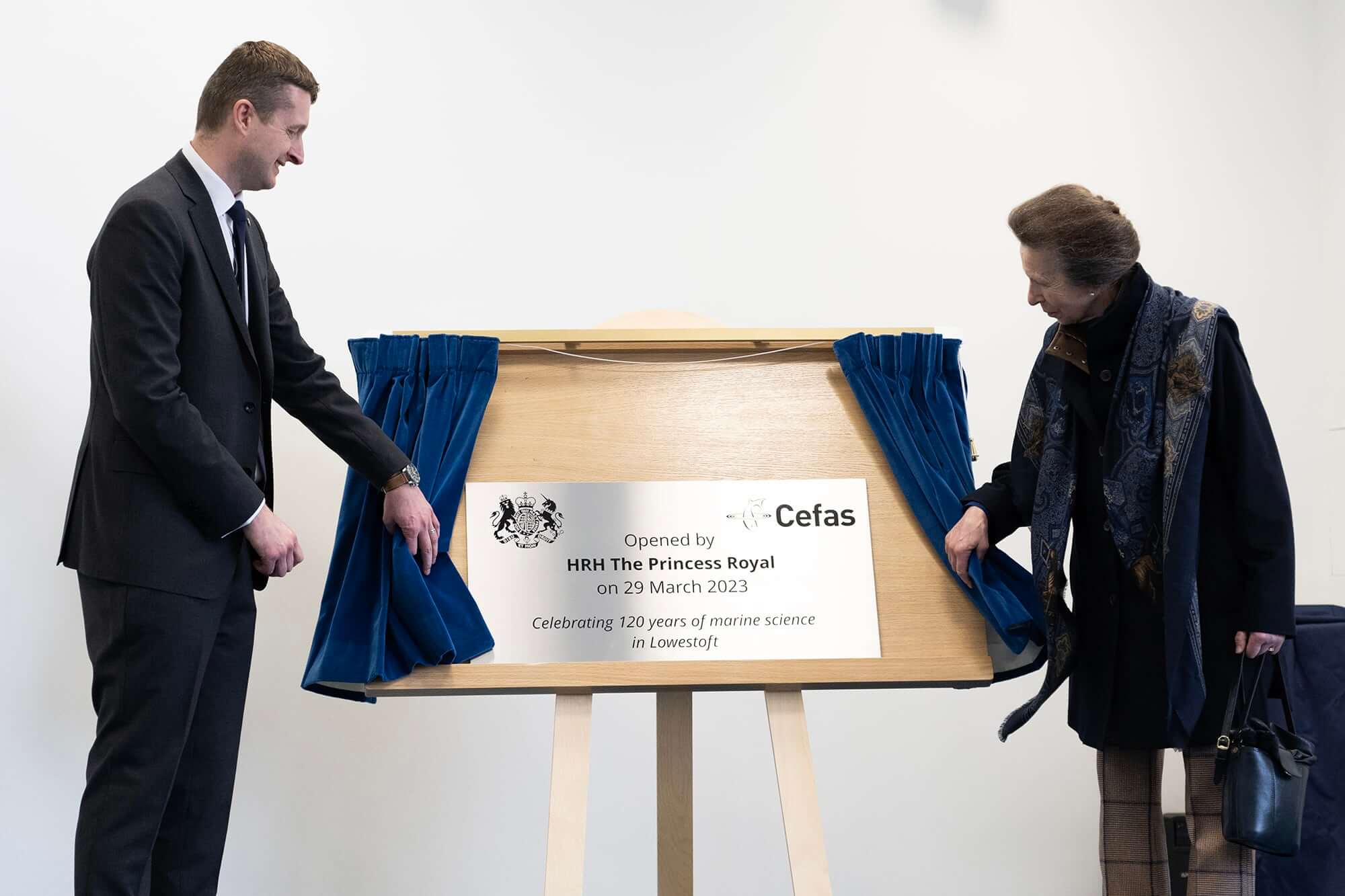 A man and a woman unveil a silver plaque for Cefas from behind blue curtains.
