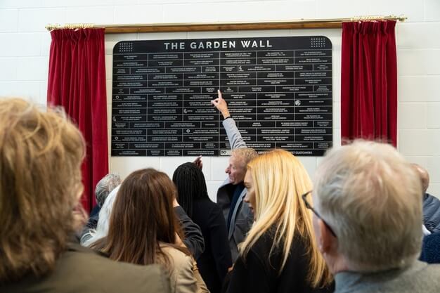 People looking and pointing at a large plaque titled "The Garden Wall" framed by red curtains.