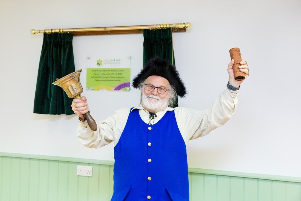 A man in a blue vest and black fuzzy hat rings bells in front of a plaque framed by green curtains.