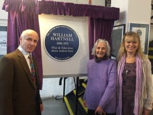 Three people pose next to a round blue plaque framed by purple curtains.