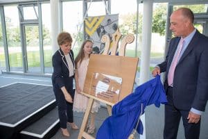 A man reveals a Perspex plaque on an easel from under a blue drape.