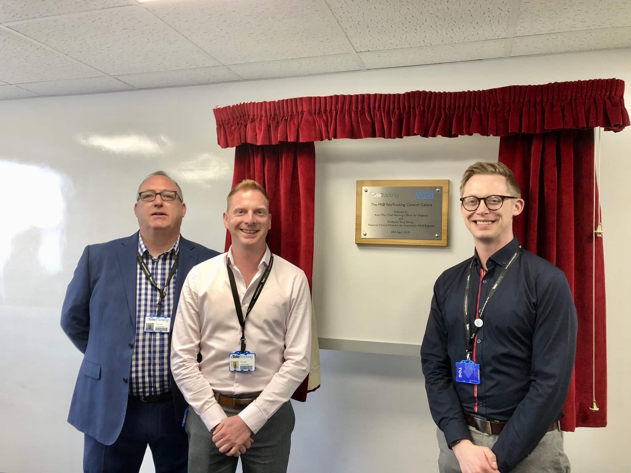 Three men in lanyards standing in front of a plaque framed by red curtains.