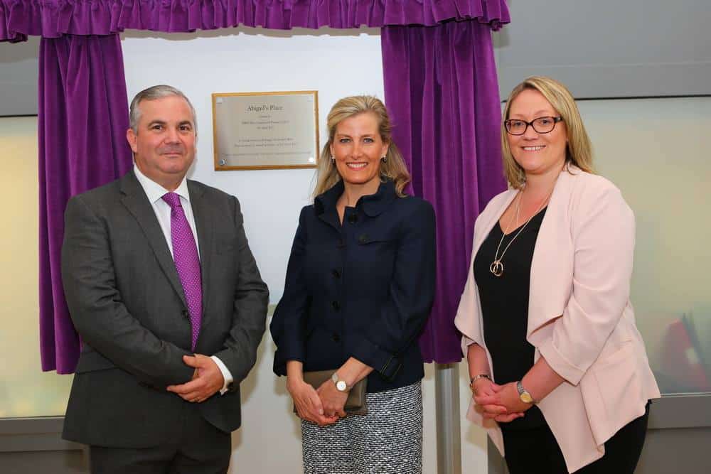 Three people stand in front of a brass plaque framed by purple curtains.