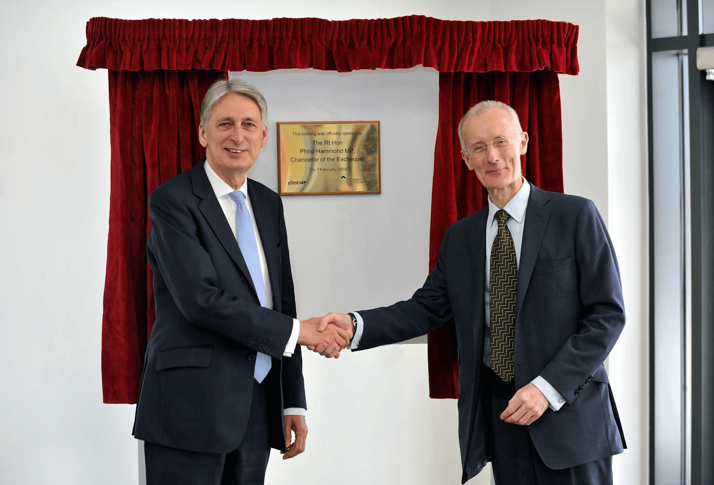 Two men shaking hands in front of a brass plaque framed by red curtains.