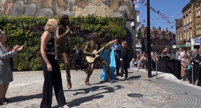 Two statues of men holding a guitar and a microphone respectively have been unveiled; a man holds the blue drape that the girl pulled off.