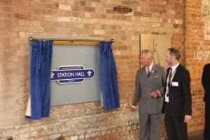 A man gestures toward a Station Hall plaque framed by blue curtains.