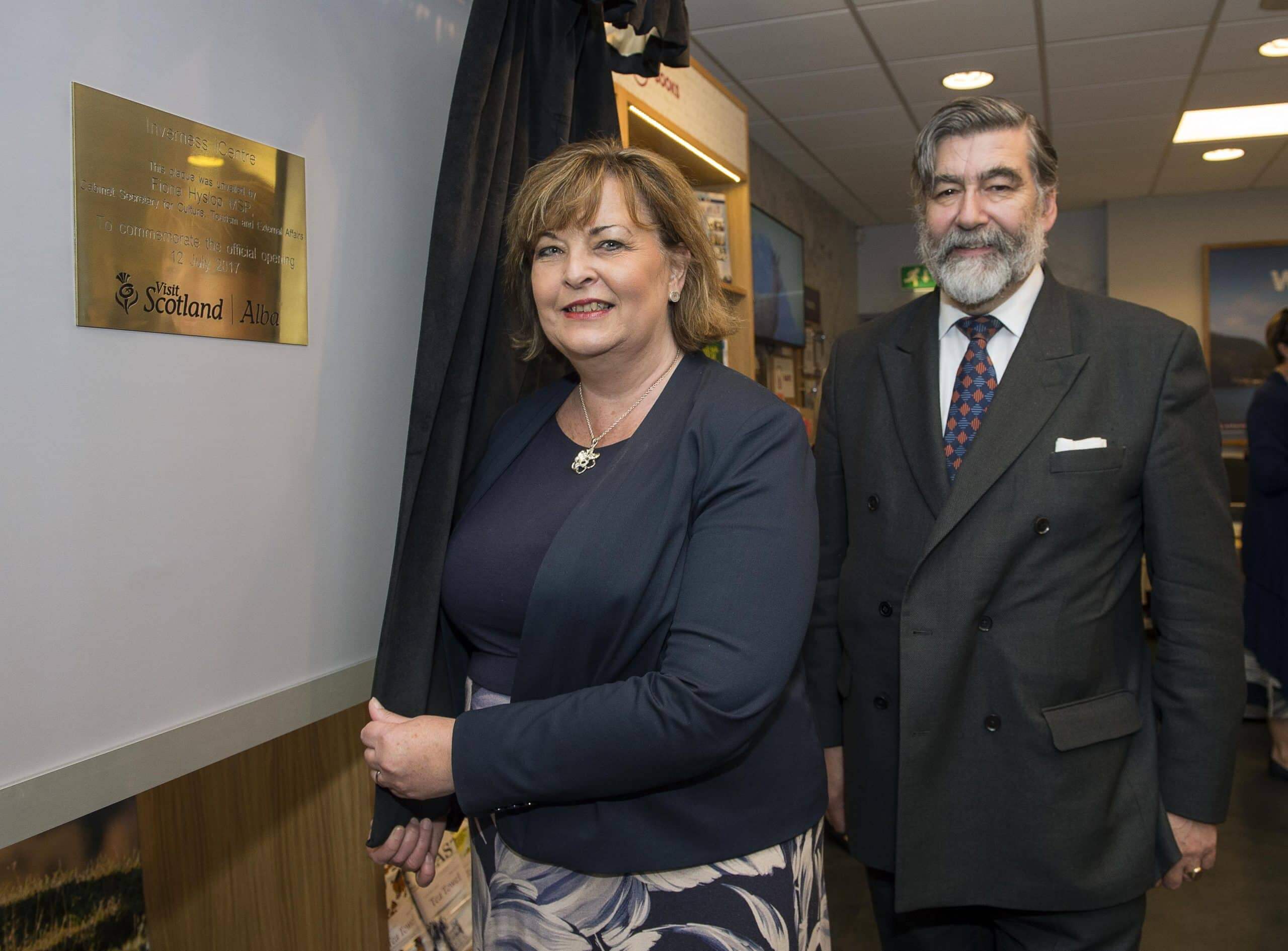 Two people stand in front of a brass plaque framed by black curtains.