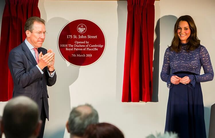 Two people standing beside a round red plaque framed by red curtains, both affixed to a wall.