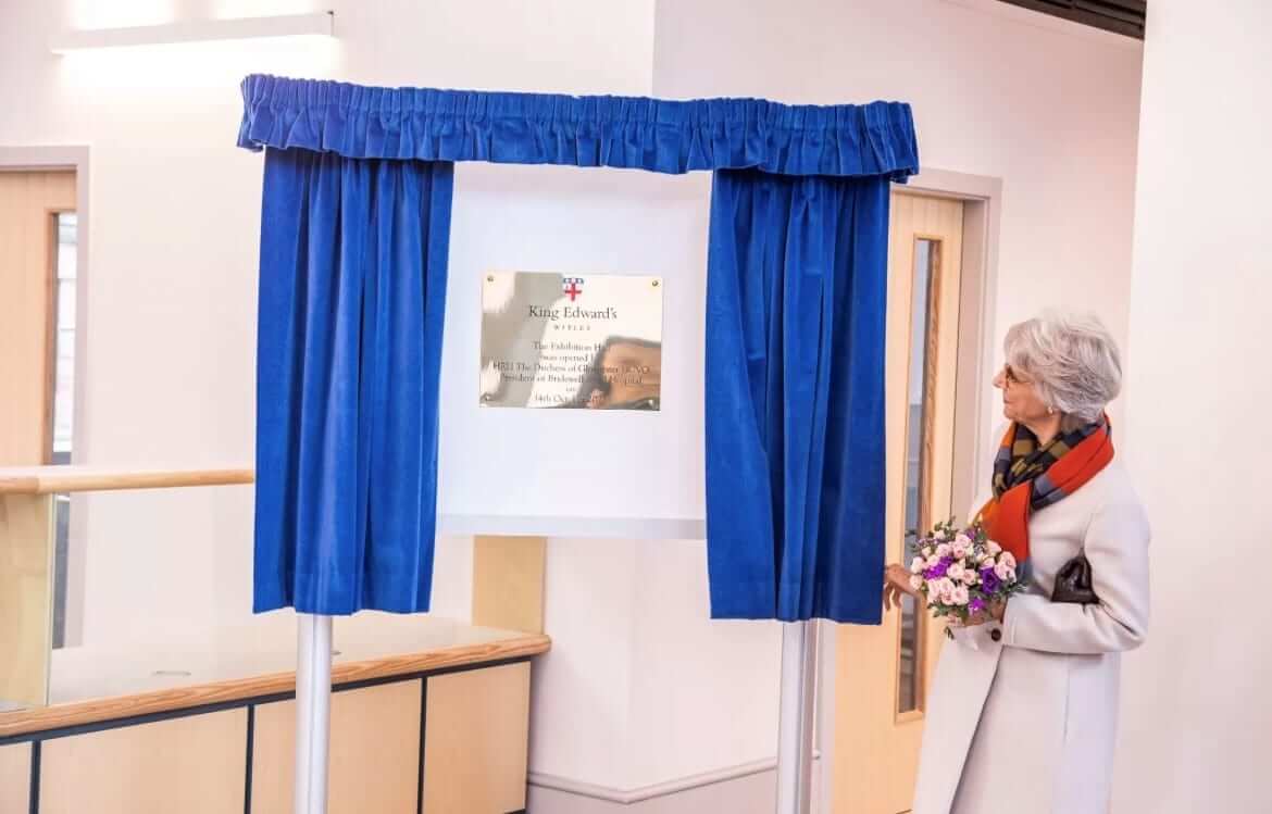 A woman looks at a brass plaque framed by blue curtains. The plaque reads in part "King Edward's."