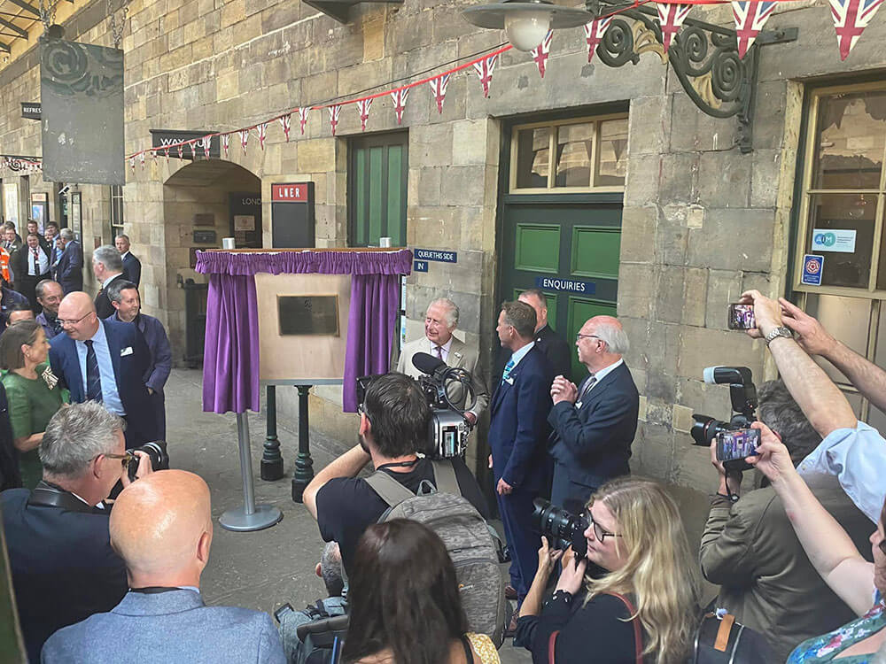 A crowd of people, some with cameras, standing around a plaque framed by purple curtains.