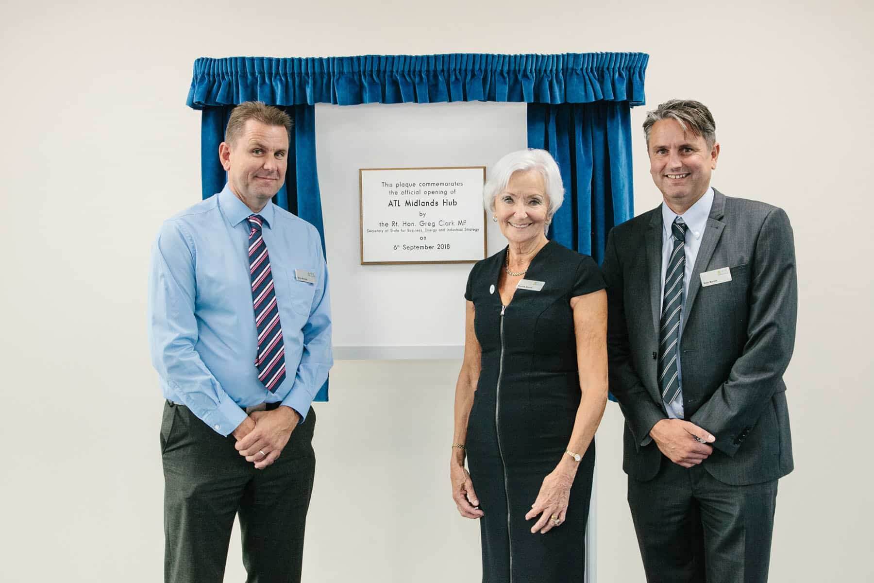 Three people standing in front of a silver plaque framed by blue curtains. The plaque commemorates the official opening of ATL Midlands Hub.