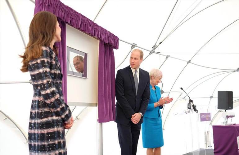 Three people standing next to a freestanding plaque framed by purple curtains. The man's face is reflected in the plaque.