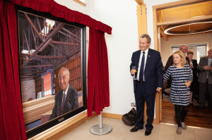 Two people look at a portrait of a smiling man in a warehouse framed by red curtains.