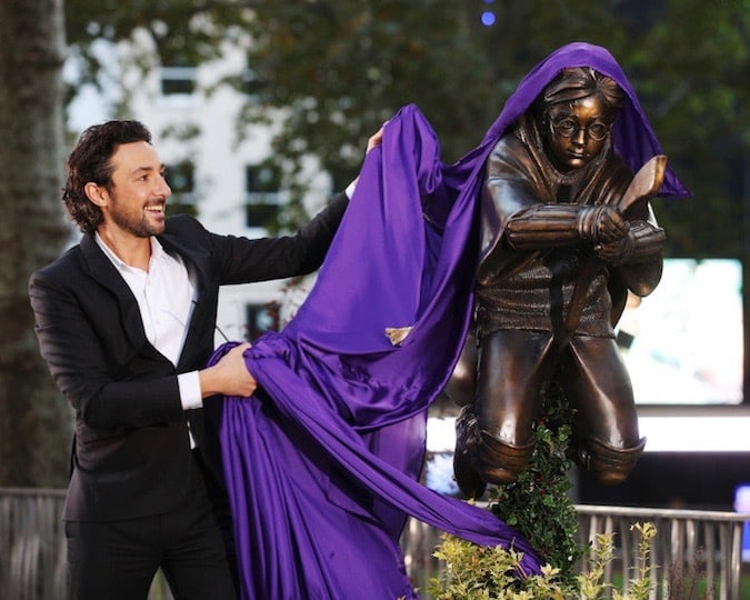 A man pulls a purple drape off a statue of a boy on a broom.