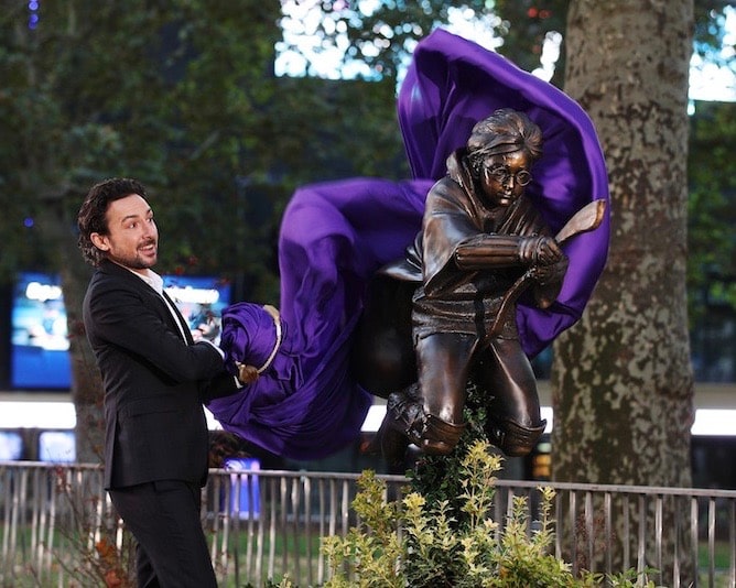 A man pulls a purple drape off a statue of a boy on a broom.