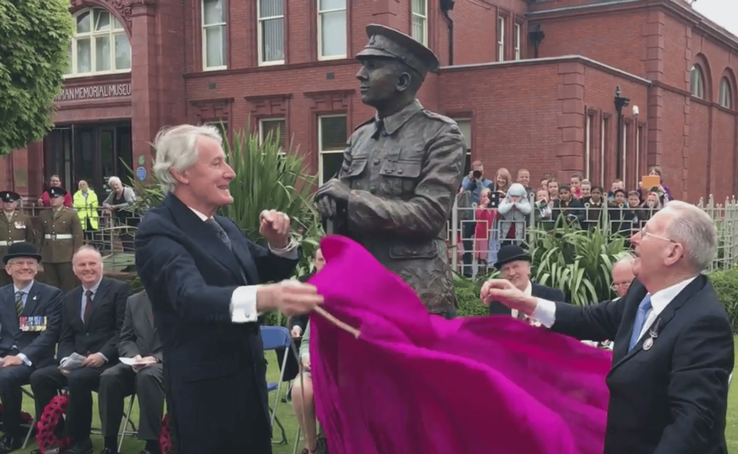 Two men unveiling a statue of a man in uniform from under a pink drape.