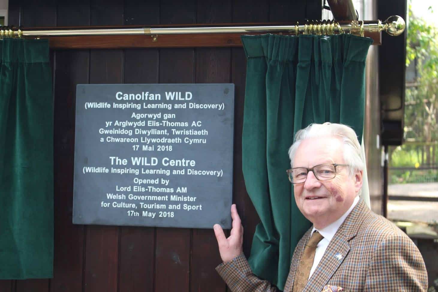 A man shows off a plaque in English and Welsh framed by green curtains.