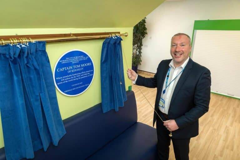 A man unveils a round blue plaque honoring Captain Tom Moore of Keighley from behind blue curtains.